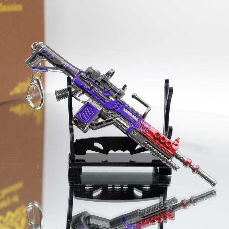 Puede incluir: Un rifle en miniatura con un esquema de color p&uacute;rpura y rojo, con una mira telesc&oacute;pica y un llavero. El arma de juguete se exhibe sobre un soporte negro, con una superficie reflectante debajo.