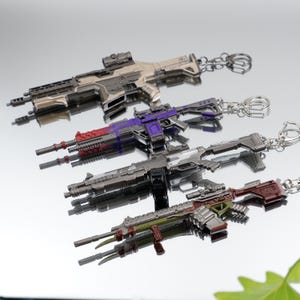 Puede incluir: Cuatro llaveros detallados con forma de pistola en varios colores: beige, morado, plateado y verde con detalles rojos. Cada llavero tiene una cadena y un cierre plateados. Los llaveros se exhiben sobre una superficie reflectante.