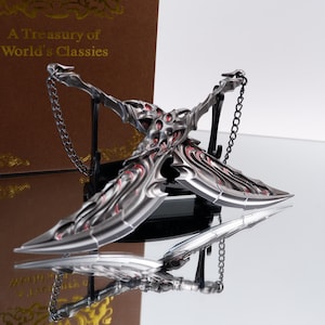 May include: A silver-colored, stylized cross-shaped pendant with red accents and a chain. The pendant is displayed on a reflective surface, with the text "A Treasury of World's Classies" visible in the background.