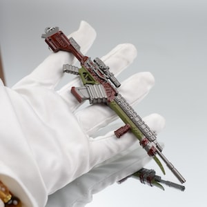 Puede incluir: Un rifle en miniatura detallado, predominantemente verde, marr&oacute;n y plateado, se sostiene en una mano enguantada. El juguete presenta una mira telesc&oacute;pica y detalles intrincados, con un acabado met&aacute;lico, lo que sugiere un art&iacute;culo de colecci&oacute;n.