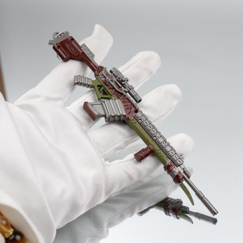 Puede incluir: Un rifle en miniatura detallado, predominantemente verde, marr&oacute;n y plateado, se sostiene en una mano enguantada. El juguete presenta una mira telesc&oacute;pica y detalles intrincados, con un acabado met&aacute;lico, lo que sugiere un art&iacute;culo de colecci&oacute;n.