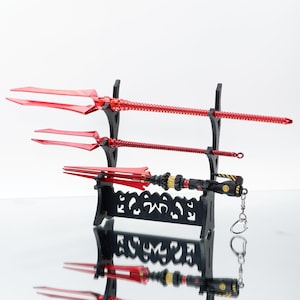 May include: Three red trident-shaped weapons displayed on a black stand. The top two tridents have long, slender shafts, while the bottom one is shorter with a detailed handle and a keychain attachment. The stand has a decorative, openwork design.
