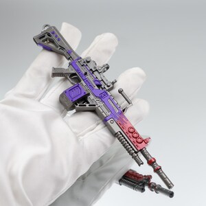 Puede incluir: Un rifle de juguete en miniatura con una combinaci&oacute;n de colores gris y morado. El arma de juguete tiene un dise&ntilde;o detallado con detalles en rojo en el ca&ntilde;&oacute;n y un peque&ntilde;o accesorio. El rifle se sostiene en una mano enguantada de blanco.