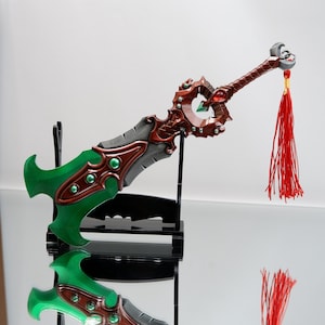 May include: A decorative fantasy sword with a green and gray blade, accented with brown and red details. The handle is brown with a silver pommel and a red tassel. The sword is displayed on a black stand.