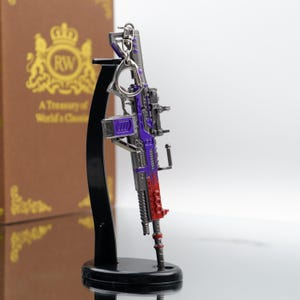 Puede incluir: Una r&eacute;plica en miniatura y detallada de un rifle, con un esquema de color p&uacute;rpura y rojo. El modelo incluye un llavero y est&aacute; montado en un soporte de exhibici&oacute;n negro. El dise&ntilde;o incorpora detalles intrincados, lo que sugiere un art&iacute;culo de colecci&oacute;n o novedad.