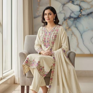 Może przedstawiać: Kremowy haftowany zestaw salwar kameez. Strój posiada kwiatowe hafty w kolorze różowym, fioletowym i zielonym. Zestaw zawiera top z długim rękawem, pasujące spodnie i drapowaną chustę. Kobieta siedzi na krześle.