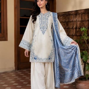 May include: An off-white long-sleeved tunic and matching wide-leg trousers featuring intricate blue floral embroidery along the neckline, cuffs, and hem. A coordinating blue patterned shawl with a scalloped lace edge drapes over one shoulder.