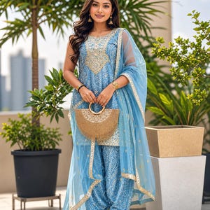 Puede incluir: Un conjunto salwar kameez bordado en azul claro con una dupatta a juego. El atuendo presenta detalles dorados y se combina con sandalias de plataforma doradas y un clutch dorado. El conjunto se usa al aire libre, con vegetación de fondo.