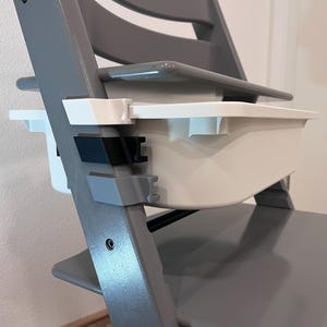 Könnte beinhalten: Ein grauer Hochstuhl mit weißem Tablett und Sitz. Der Stuhl hat ein modernes Design mit einem stabilen Rahmen und verstellbaren Funktionen. Das Tablett ist mit schwarzen und weißen Halterungen am Stuhl befestigt.