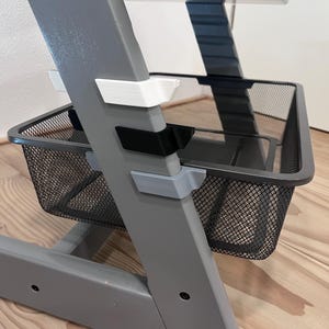 May include: A gray high chair with a black wire mesh basket. The chair has white, black, and gray accents. The chair is on a light brown wooden floor.