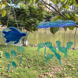 Puede incluir: Dos móviles colgantes con temática marina. Uno presenta una tortuga marina azul, el otro un diseño de olas azules. Ambos tienen detalles colgantes en forma de hojas y peces en tonos verdes y azules. Los móviles están suspendidos de una rama de árbol.