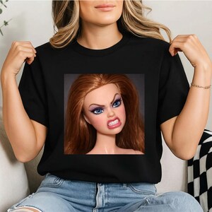 以下が含まれることがあります： 怒った表情の人形がプリントされた黒いTシャツ。人形は赤い髪と青い目をしています。Tシャツはクルーネックで、着用されています。