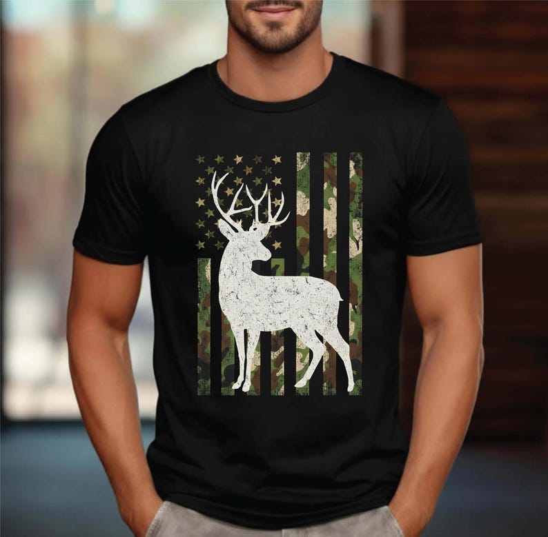 May include: Black t-shirt featuring a white silhouette of a deer with large antlers. The deer is set against a camouflage pattern resembling the American flag. The shirt has a crew neck and short sleeves.