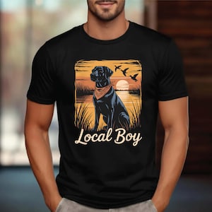 May include: Black t-shirt featuring a graphic of a black dog wearing a bandana, set against a sunset scene with birds. The text "Local Boy" is displayed below the image. The shirt is a classic crew neck.