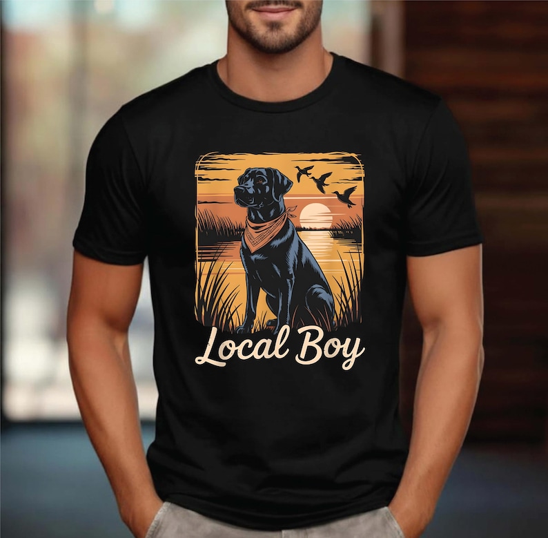 May include: Black t-shirt featuring a graphic of a black dog wearing a bandana, set against a sunset scene with birds. The text "Local Boy" is displayed below the image. The shirt is a classic crew neck.