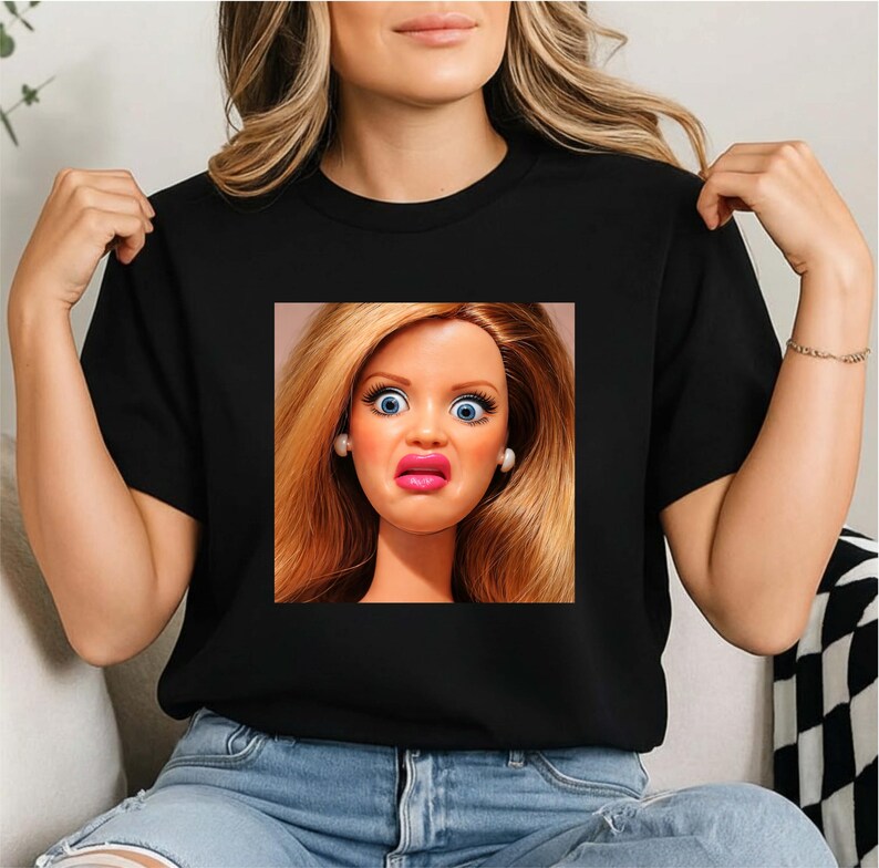 以下が含まれることがあります： 黒いTシャツ。驚いた表情で青い目を大きく見開いた人形の顔がプリントされています。人形はブロンドの髪とピンクの唇をしています。Tシャツはクルーネックです。