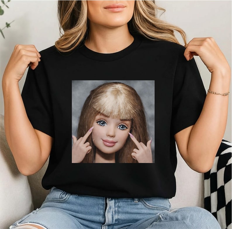 以下が含まれることがあります： 黒いTシャツ。ブロンドの髪、青い目、ピンクの爪をした人形の正方形の画像が特徴です。人形は両手で2本の指を立てています。Tシャツはクルーネックです。