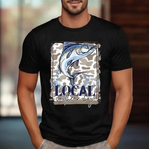 May include: Black t-shirt featuring a detailed illustration of a fish in shades of blue and white, set against a camouflage background. The text "LOCAL southern boy" is displayed below the fish in a stylized font.