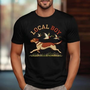 May include: Black t-shirt with an embroidered design featuring a running dog, ducks, and the text "LOCAL BOY" in orange and red. The dog is brown and white, and the ducks are green and white. The design is set against a green grass background.