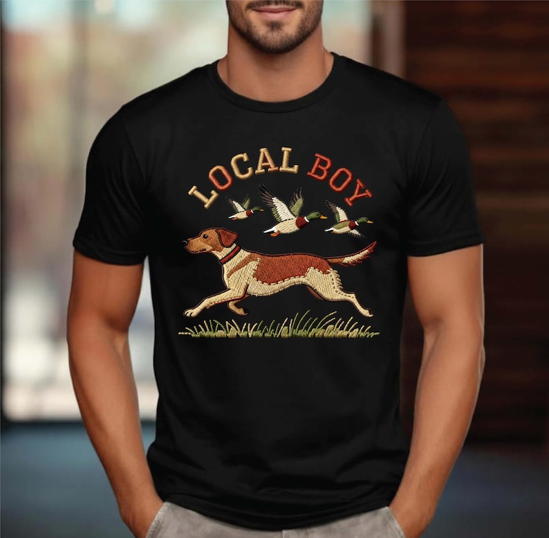 May include: Black t-shirt with an embroidered design featuring a running dog, ducks, and the text "LOCAL BOY" in orange and red. The dog is brown and white, and the ducks are green and white. The design is set against a green grass background.