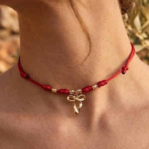 Può includere: Collana con cordino rosso con dettagli dorati e un ciondolo a forma di fiocco. La collana presenta piccole perline dorate e un delicato ciondolo a fiocco, creando un design semplice ma elegante. Un accessorio elegante.