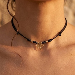 Può includere: Una collana girocollo nera con un ciondolo a forma di fiocco color oro. La collana è realizzata con un cordoncino sottile e intrecciato. Il ciondolo a forma di fiocco è centrato sulla collana ed è un elemento decorativo delicato. La collana è indossata intorno al collo.