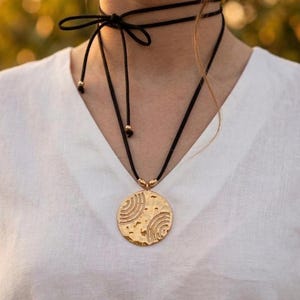 Op de afbeelding: Een zwarte choker-ketting met een gouden hanger. De hanger is een groot, gestructureerd, cirkelvormig ontwerp met gebogen lijnen. De ketting wordt met een strik in de nek vastgemaakt en wordt gedragen over een wit V-hals shirt.