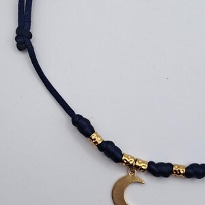 May include: A navy blue cord necklace featuring a gold-toned crescent moon charm. The cord is accented with small, textured gold-toned cylindrical beads positioned between knots, creating a structured design along the length of the necklace.
