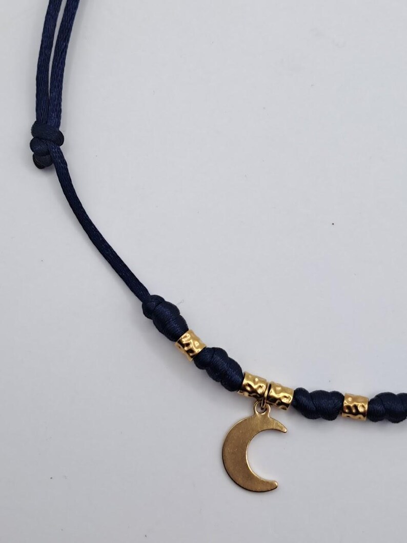 May include: A navy blue cord necklace featuring a gold-toned crescent moon charm. The cord is accented with small, textured gold-toned cylindrical beads positioned between knots, creating a structured design along the length of the necklace.