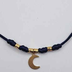 May include: A necklace featuring a dark blue cord with knotted sections and three small, textured gold-toned metal beads. A smooth, gold-toned crescent moon charm hangs from the center of the cord, suspended by a small circular metal ring.