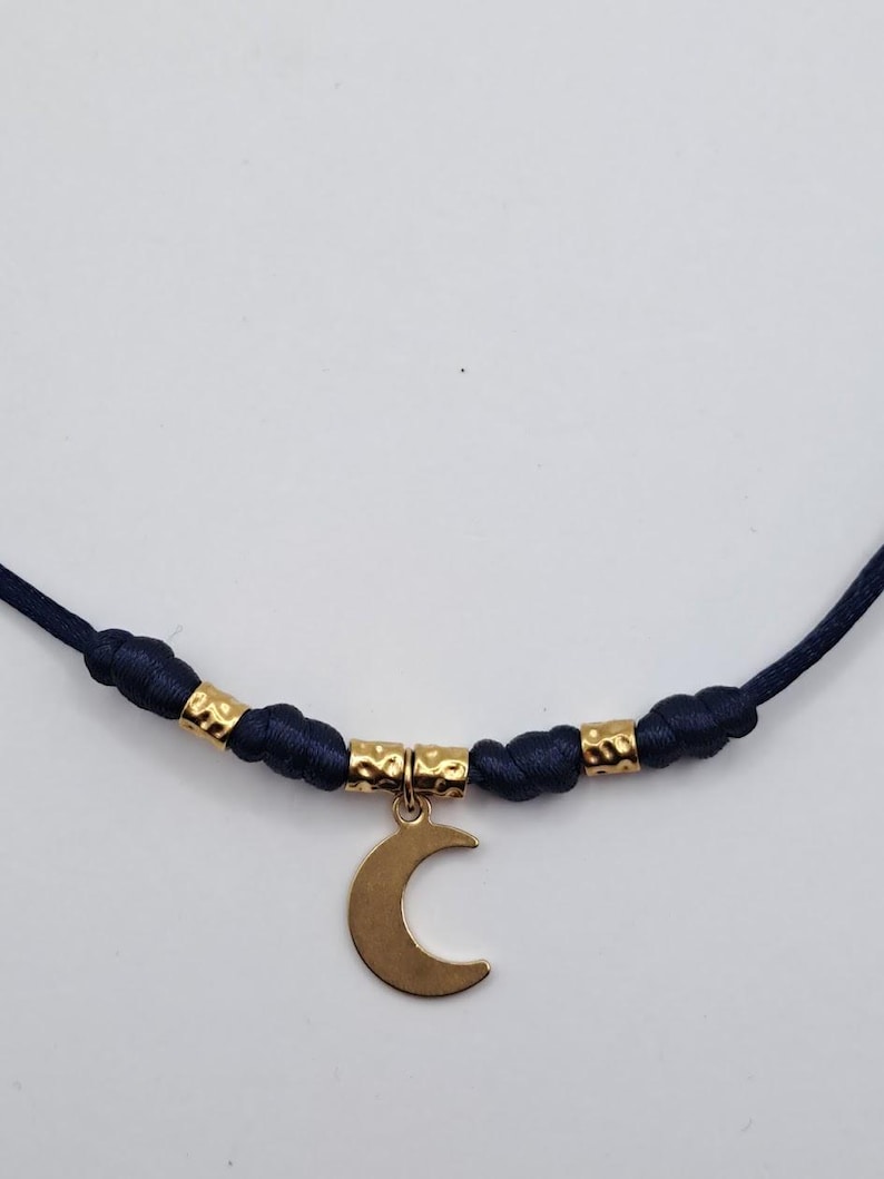 May include: A necklace featuring a dark blue cord with knotted sections and three small, textured gold-toned metal beads. A smooth, gold-toned crescent moon charm hangs from the center of the cord, suspended by a small circular metal ring.