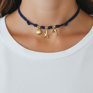 Può includere: Una collana girocollo blu navy con ciondoli color oro. I ciondoli includono una conchiglia, una stella marina e un delfino. La collana è indossata attorno al collo, su una camicia bianca.