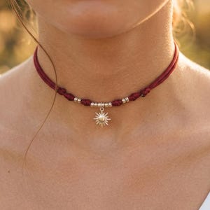 Può includere: Una collana girocollo bordeaux con un ciondolo a forma di sole argentato. La collana presenta un design a cordoncino intrecciato con perline d'argento. Il ciondolo a forma di sole è dettagliato con raggi che si estendono verso l'esterno.