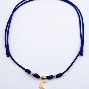 May include: A necklace featuring a thin, dark blue cord with adjustable knots. The center of the cord is adorned with two small, textured gold-toned metal beads flanking a central gold-toned crescent moon pendant that hangs downward.