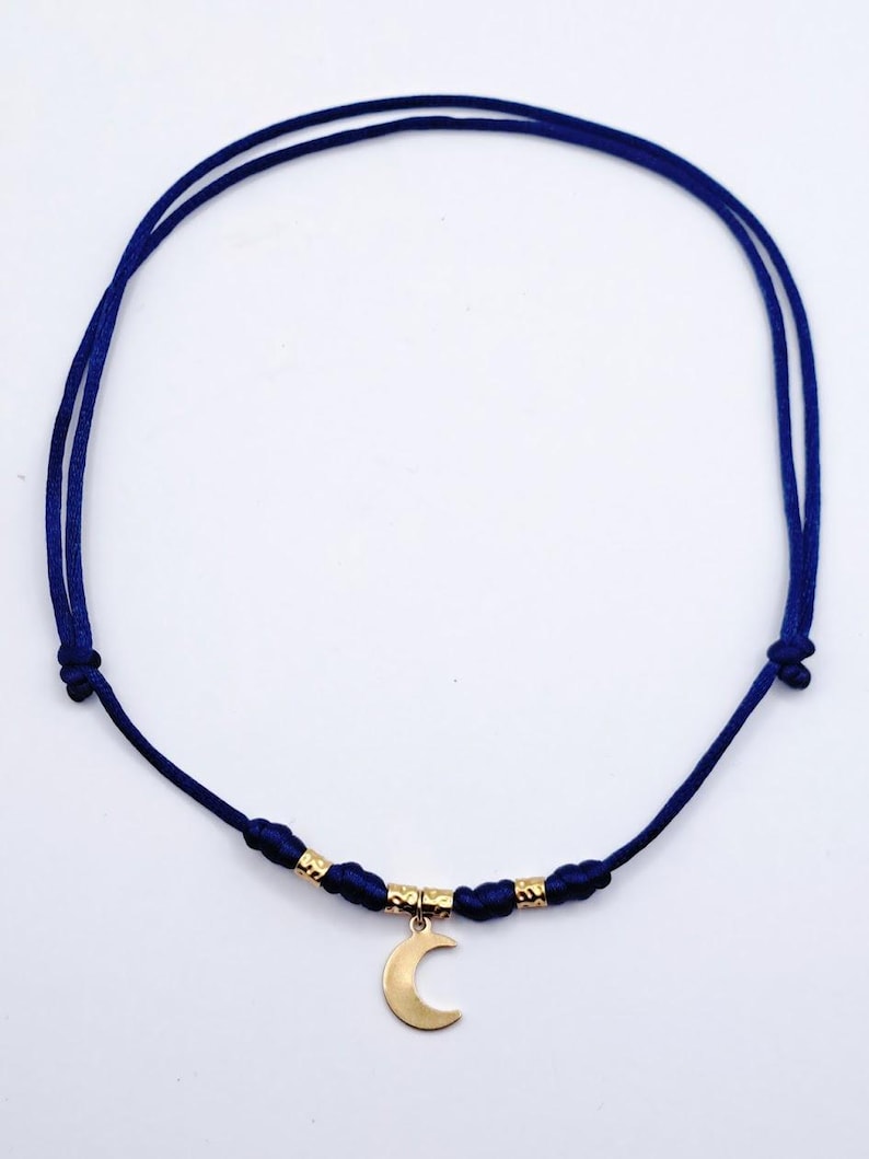 May include: A necklace featuring a thin, dark blue cord with adjustable knots. The center of the cord is adorned with two small, textured gold-toned metal beads flanking a central gold-toned crescent moon pendant that hangs downward.