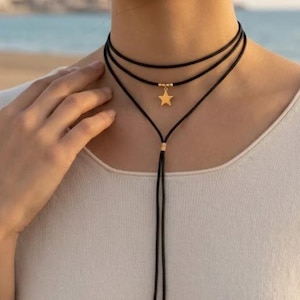 Puede incluir: Collar gargantilla negro con un colgante de estrella dorada. El collar tiene tres hebras y una corbata larga y ajustable. El colgante de estrella es de color dorado. El collar se lleva alrededor del cuello.