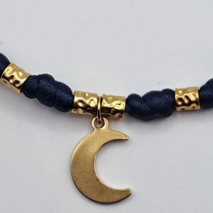 May include: A necklace featuring a dark blue cord with a series of gold-toned, hammered-texture cylindrical beads. A smooth, flat gold-toned crescent moon charm hangs from the center of the cord, suspended by a small circular jump ring.