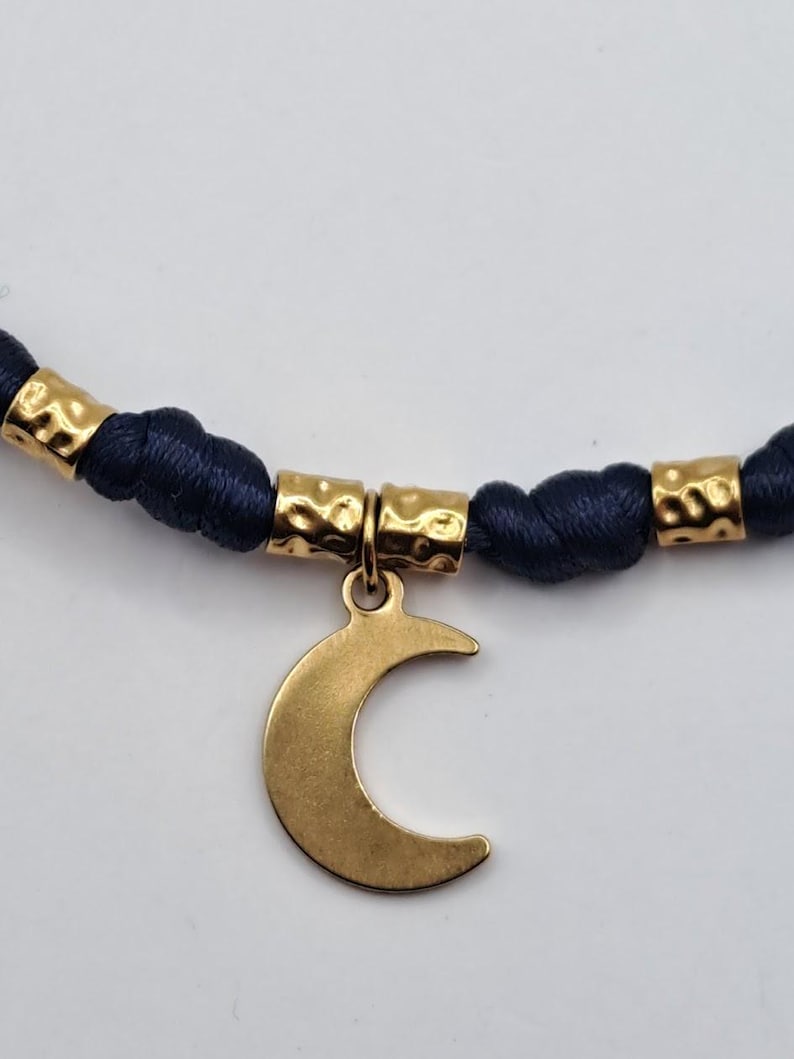 May include: A necklace featuring a dark blue cord with a series of gold-toned, hammered-texture cylindrical beads. A smooth, flat gold-toned crescent moon charm hangs from the center of the cord, suspended by a small circular jump ring.