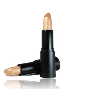 May include: A lipstick with a shimmering champagne-colored bullet housed in a matte black cylindrical tube. One lipstick stands upright while another lies horizontally at its base, both featuring a metallic finish and a smooth, slanted tip.