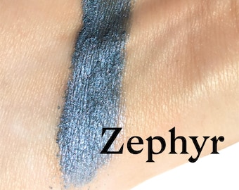 May include: A swatch of metallic blue eyeshadow with a shimmering finish applied to skin. The word Zephyr is printed in a black serif font next to the blue pigment, which displays a cool-toned, reflective texture with visible fine particles.