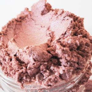May include: A close-up of a jar of loose, shimmery, pink eyeshadow. The eyeshadow is a light pink with a subtle shimmer.