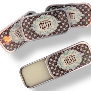 Op de afbeelding: Vier kleine zilveren blikjes met een bruin en wit bloemenpatroon. De blikjes hebben een etiket met de tekst "Crush Velvet Solid Perfume". Eén blikje is open en laat een witte balsem zien.