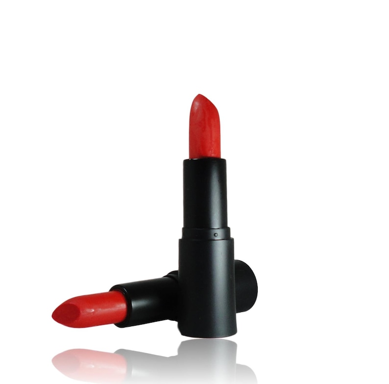 May include: A vibrant red lipstick in a matte black cylindrical tube. One lipstick stands upright with the cap removed, revealing the bright red pigment, while a second identical lipstick lies horizontally at its base on a white surface.