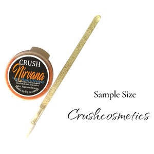 Puede incluir: Un peque&ntilde;o recipiente redondo de maquillaje mineral vegano Crush Nirvana con etiqueta negra y texto naranja, acompa&ntilde;ado de un pincel aplicador largo y fino cubierto de purpurina dorada. El texto Sample Size y Crushcosmetics aparece en negro.