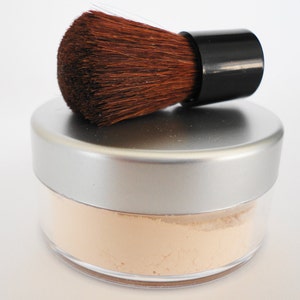 May include: A silver jar with a clear lid containing a light beige powder. A brown makeup brush with a black handle is on top of the jar.