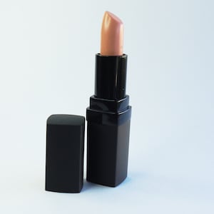 May include: A lipstick with a light beige, nude-toned pigment extended from a square, matte black casing. The matching black cap is positioned upright next to the base of the lipstick tube, which features a smooth, angular design.