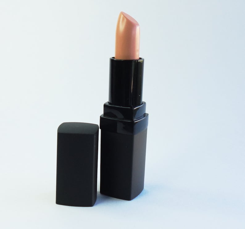 May include: A lipstick with a light beige, nude-toned pigment extended from a square, matte black casing. The matching black cap is positioned upright next to the base of the lipstick tube, which features a smooth, angular design.