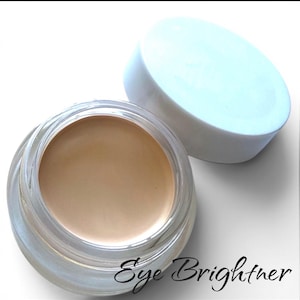 May include: A small, round, clear glass jar filled with a smooth, beige-toned cream cosmetic product. The jar has a white, circular lid placed beside it. The text "Eye Brightner" is printed in a black, cursive script below the jar.