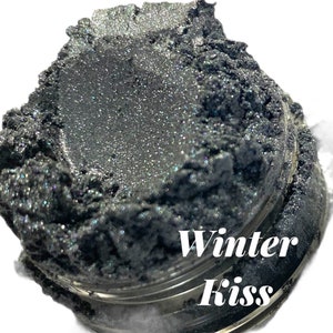 Op de afbeelding: Een close-up van een pot met donkergrijze, iriserende glitter met de tekst "Winter Kiss" erop gedrukt.