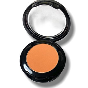 May include: Open compact of peach-colored face powder with a black case and a built-in mirror. The powder is a matte finish and is surrounded by a black rim. The compact is open, revealing the powder and mirror.